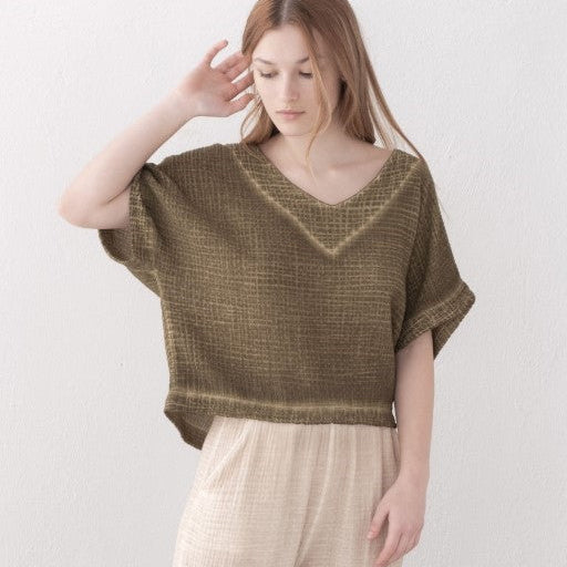 A woman wearing a textured olive green short-sleeve knit top with a V-neck and light beige wide-leg trousers, standing against a plain white wall.