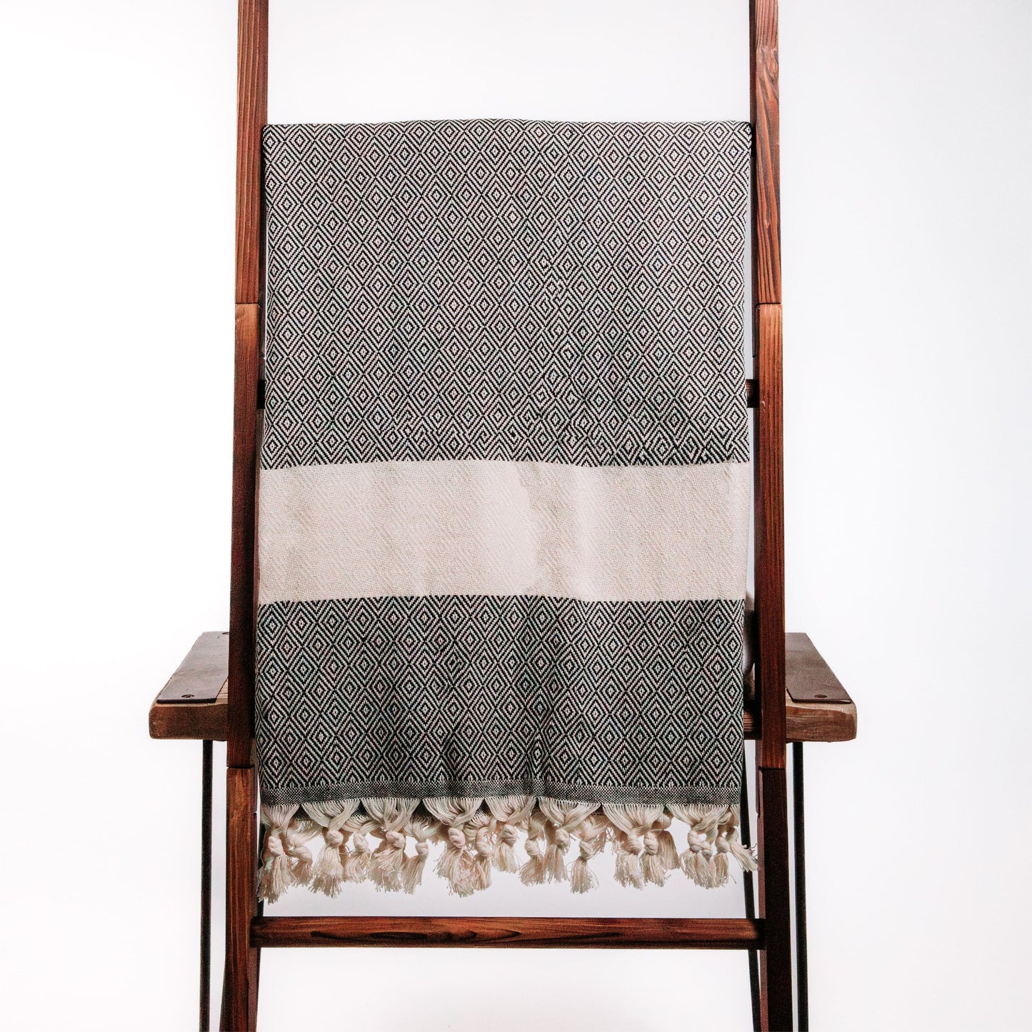 A beige and black peshtemal towel with a geometric pattern, draped over the back of a wooden chair against a white background.