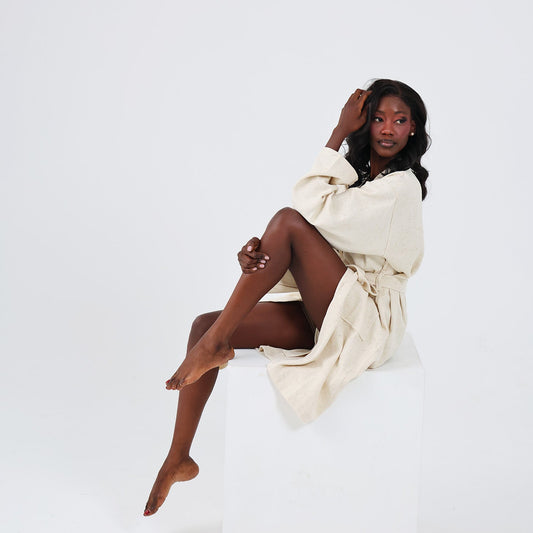 A woman posing on a white block in a cream-colored linen robe against a seamless white studio background.