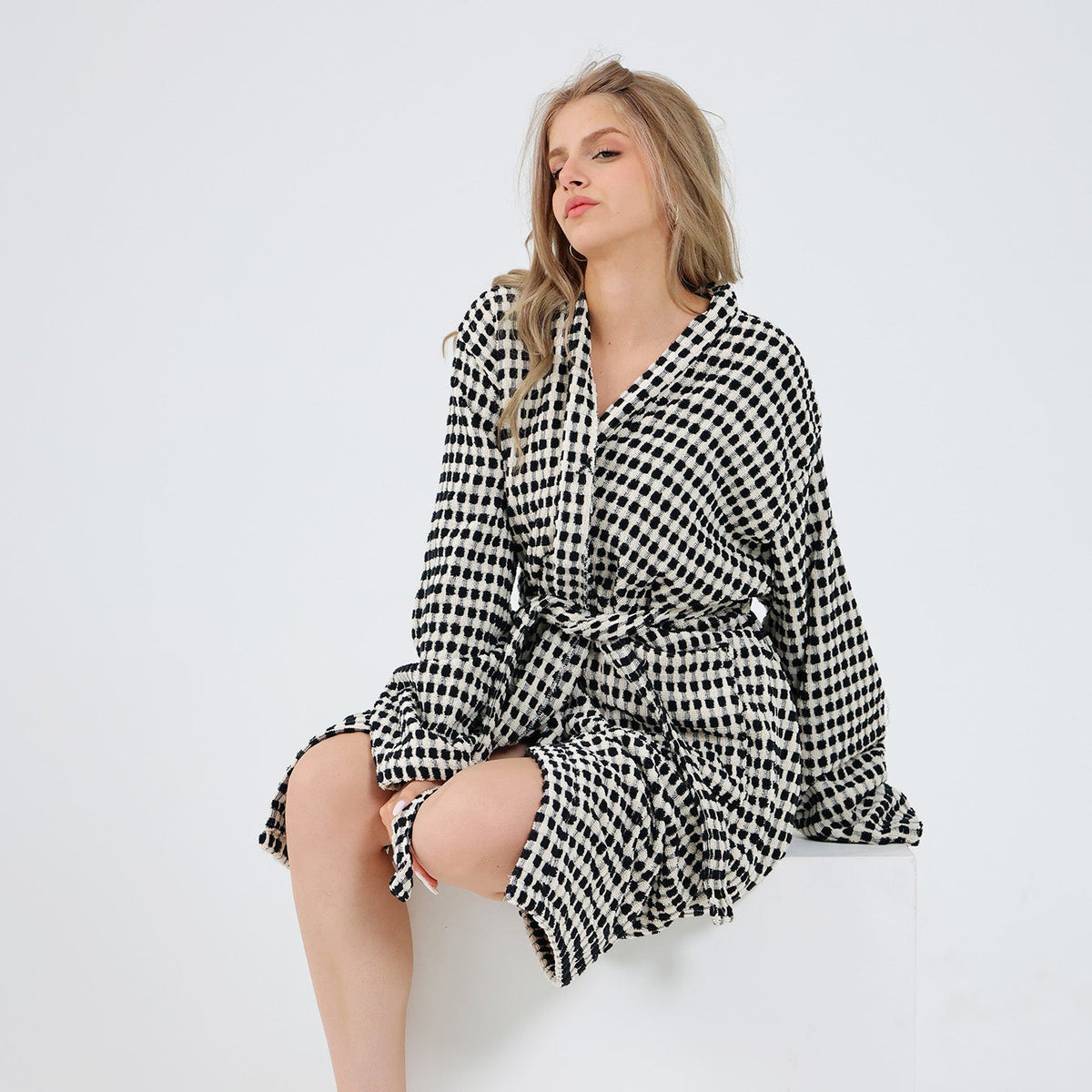 A woman sits barefoot on a white block against a plain white studio background, wearing a long-sleeve belted robe in a black and white small-scale check pattern.