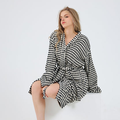 A woman sits barefoot on a white block against a plain white studio background, wearing a long-sleeve belted robe in a black and white small-scale check pattern.