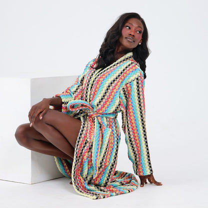 A woman wearing a long-sleeve striped robe sits on a white block against a plain white studio background. The robe features a multicolor zigzag stripe pattern with black, orange, teal, yellow, and cream tones.