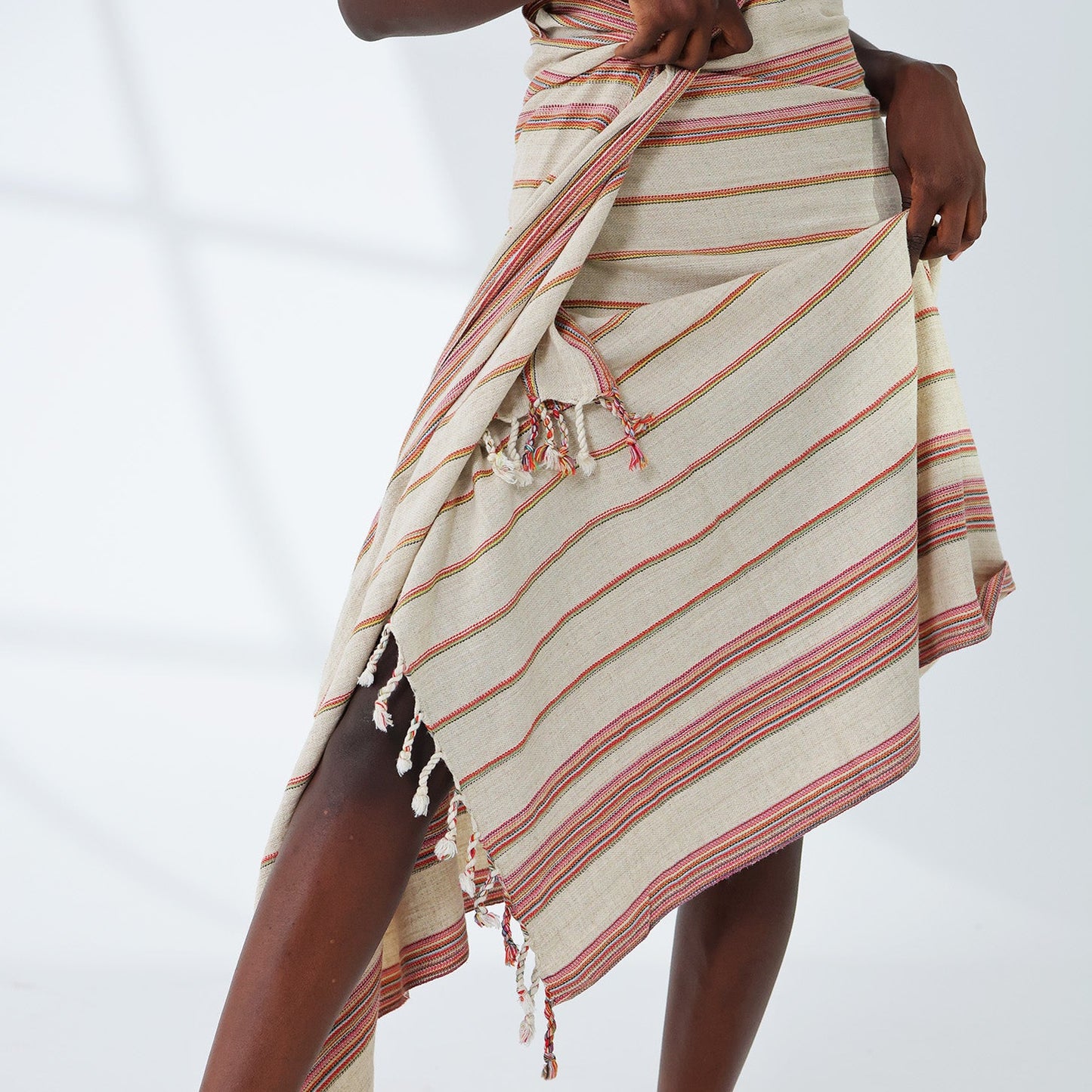 A model wearing a beige and pink striped cotton sarong wrap skirt with tassels, styled with a black crop top and black slide sandals against a white studio background.