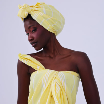 A woman wearing a pale yellow striped towel wrap as a one-shoulder dress and a matching striped towel turban headwrap, posed against a light gray studio background.