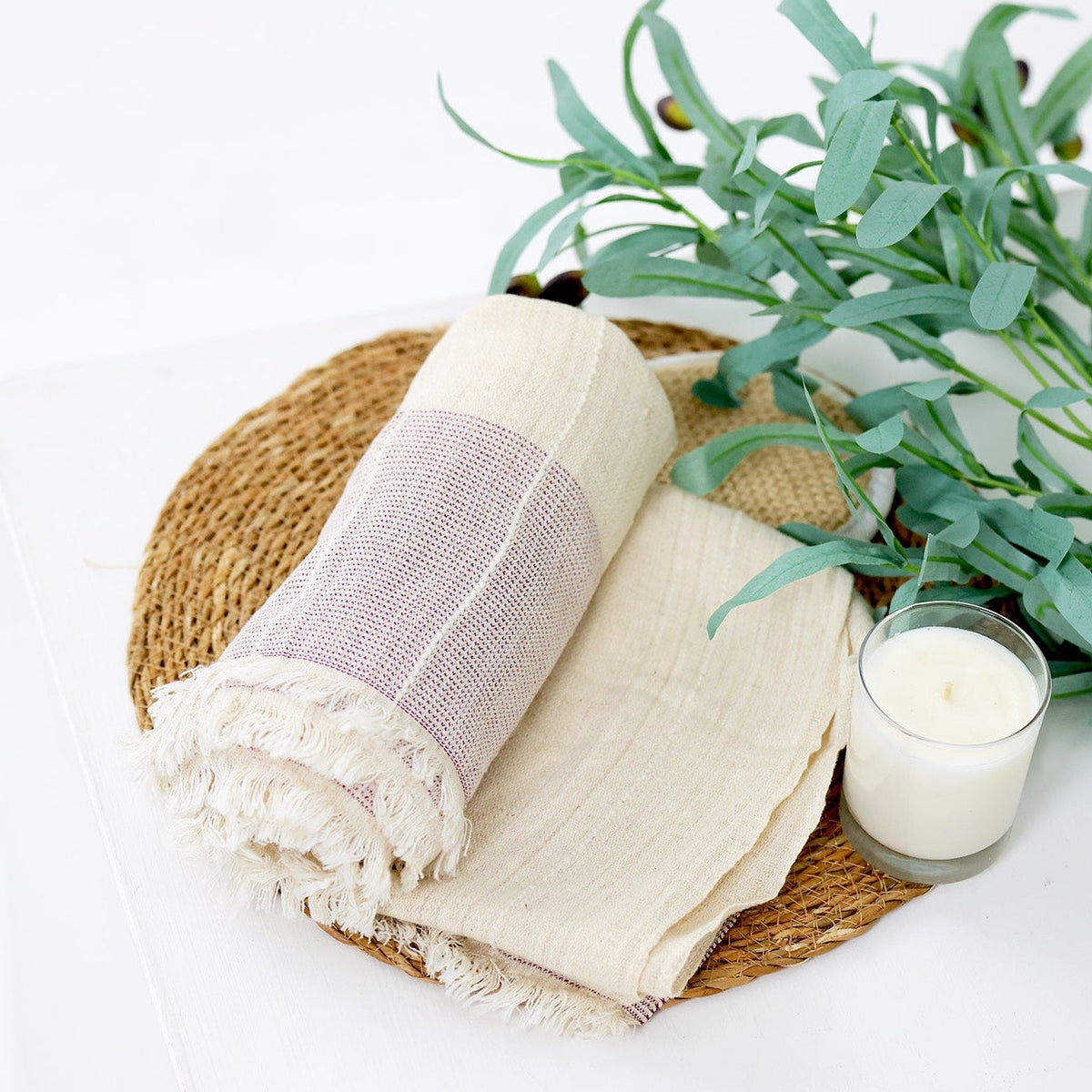 A styled flat-lay of a woven round tray holding a folded cream linen cloth and a rolled cream-and-soft pink striped textile with fringed edges, alongside a white candle and green eucalyptus stems on a white background.