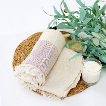 A styled flat-lay of a woven round tray holding a folded cream linen cloth and a rolled cream-and-soft pink striped textile with fringed edges, alongside a white candle and green eucalyptus stems on a white background.