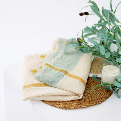A folded cream-colored cotton scarf with soft sage green and mustard yellow stripes, styled on a woven placemat with eucalyptus leaves and a candle on a white background.