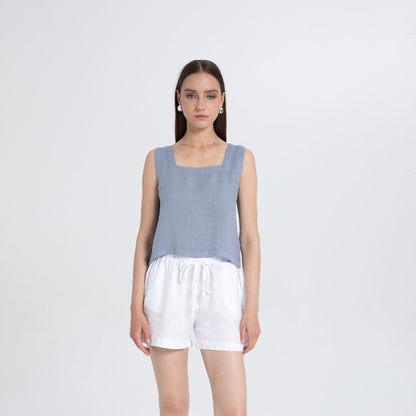 A woman stands against a light gray studio background wearing a sleeveless dusty blue top with a square neckline and white shorts.