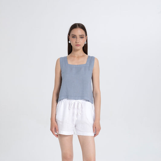 A woman stands against a light gray studio background wearing a sleeveless dusty blue top with a square neckline and white shorts.