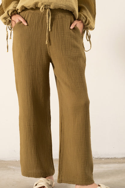 A person wearing wide-leg textured trousers in a warm camel brown, paired with a matching top and beige slide sandals, standing against a plain off-white wall.