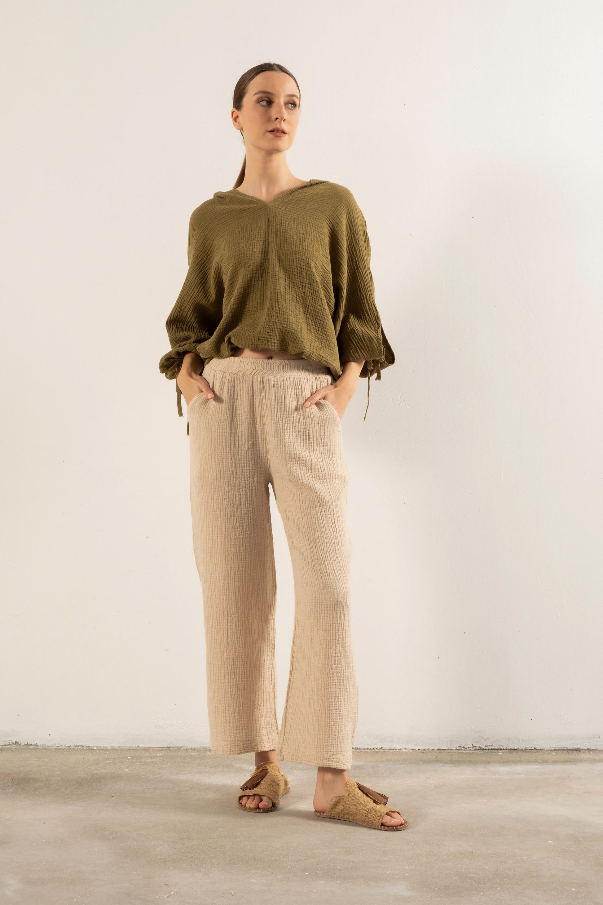 A woman stands against a plain off-white wall wearing an olive green long-sleeve top with tie details at the cuffs, beige pleated wide-leg trousers, and tan sandals.