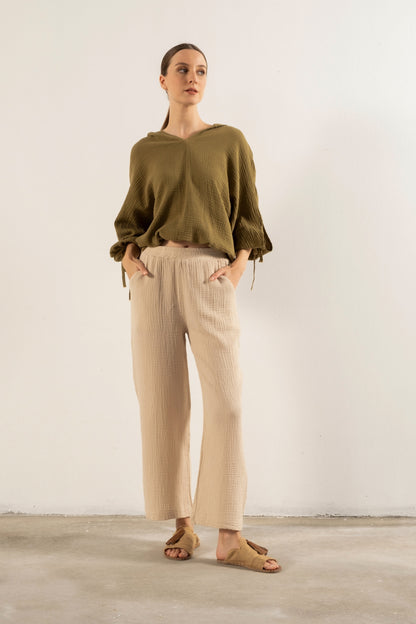A woman stands against a plain off-white wall wearing an olive green long-sleeve top with tie details at the cuffs, beige pleated wide-leg trousers, and tan sandals.