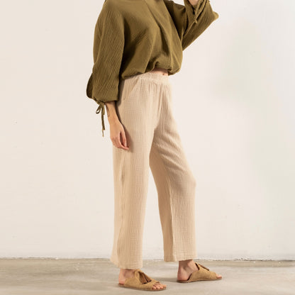 A woman modeling a loose olive green long-sleeve top and cream wide-leg trousers, standing in a minimalist studio with a light beige wall and concrete floor.