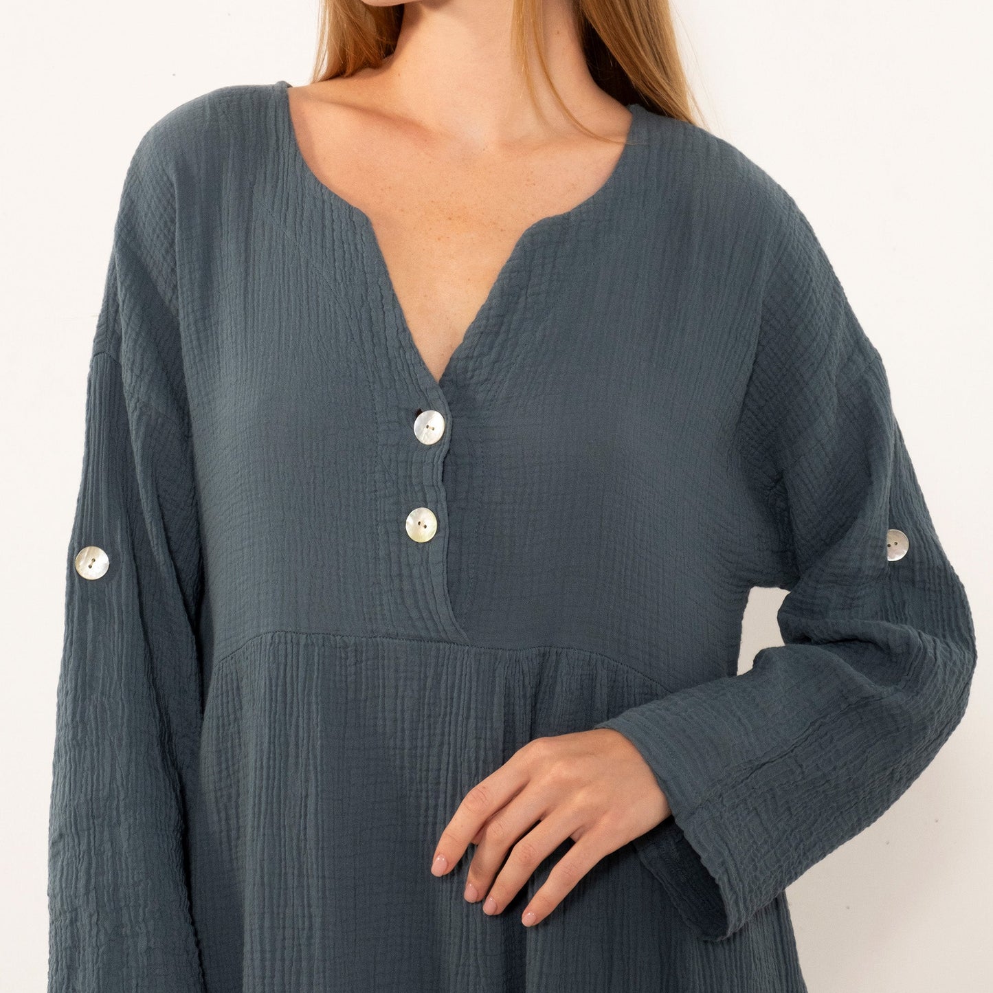 A woman wearing a dark slate blue crinkle-textured dress with a split neckline, three white buttons, and decorative white buttons on the cuffs, shown against a plain off-white background.