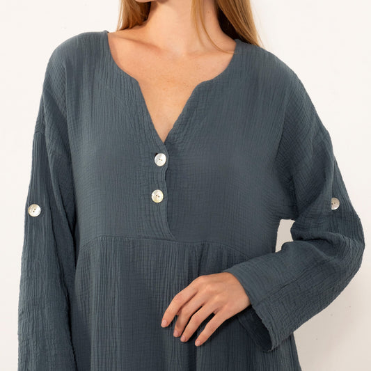 A woman wearing a dark slate blue crinkle-textured dress with a split neckline, three white buttons, and decorative white buttons on the cuffs, shown against a plain off-white background.