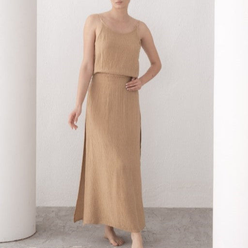 Woman wearing a beige top and long skirt standing against a white wall.