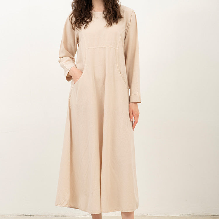 A woman modeling a long-sleeve, midi-length beige dress with side pockets, paired with brown leather slide sandals, standing against a plain off-white studio background.