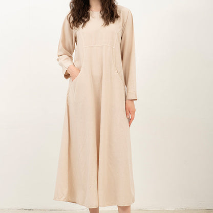 A woman modeling a long-sleeve, midi-length beige dress with side pockets, paired with brown leather slide sandals, standing against a plain off-white studio background.