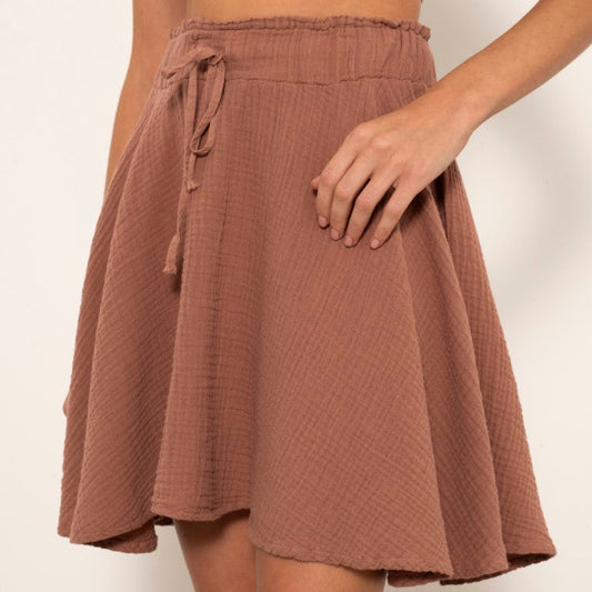 A woman wearing a dusty rose brown textured cotton gauze mini skirt with a tie waist and a matching crop top, shown against a plain off-white background.