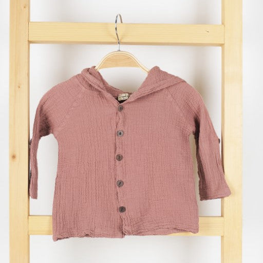 A dusty rose pink crinkle gauze button-up cardigan with a hood, hanging on a wooden hanger from a light wood ladder against a white wall.